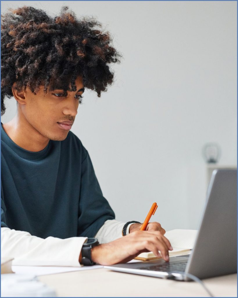 Young adult sitting at a computer screen wearing a blue and white shirt and holding a pen.